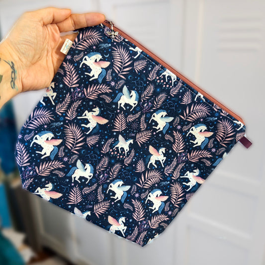 A hand holding a zippered knitting project bag with a dark blue fabric flying unicorns and plants print against a neutral background