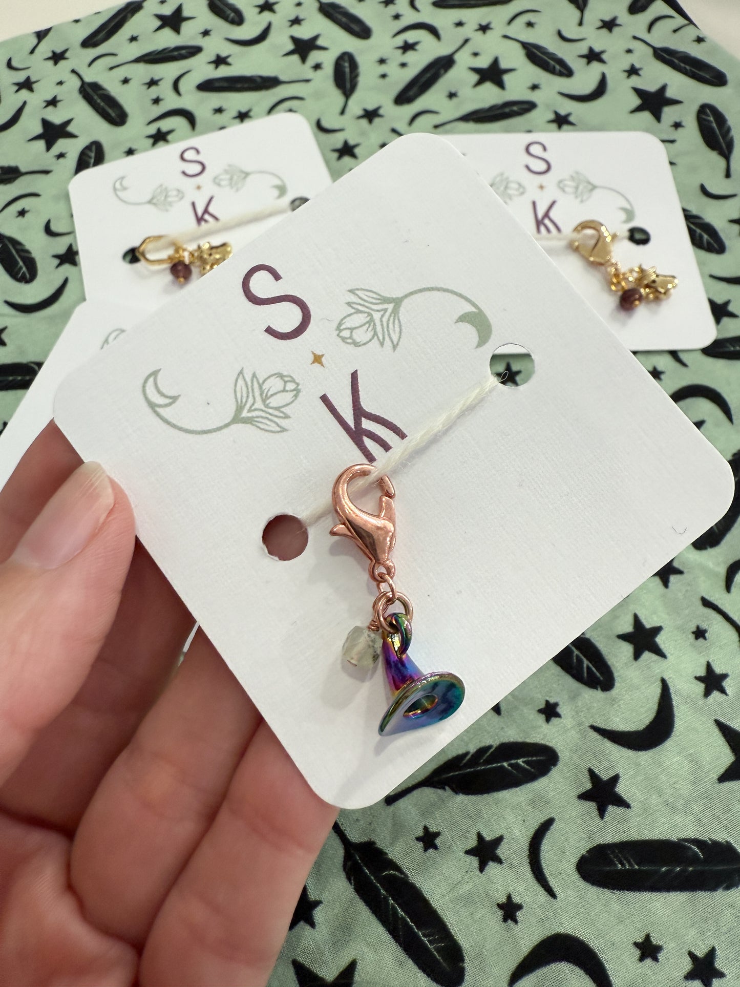 A hand holding a white card with a charm shaped like a witch hat, and other charms in the background against a mint colored fabric with feathers, moons, and stars