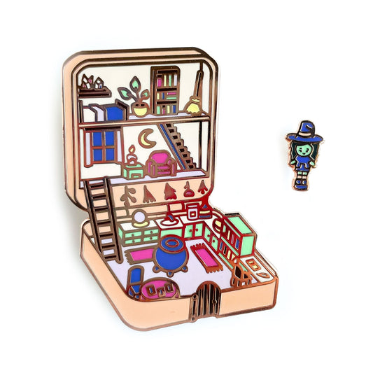 An enamel pin set that is Polly Pocket style compact of a witch house with a little witch mini doll. The compact house has a cauldron, crystal ball, tarot cards, crystals, and a broom. The compact is a pastel peach color with mint green, hot pink and purple details. 