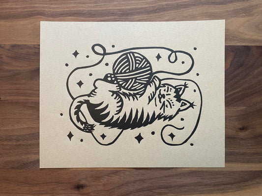 Black and white linocut of a cat playing with a ball of yarn on beige paper.