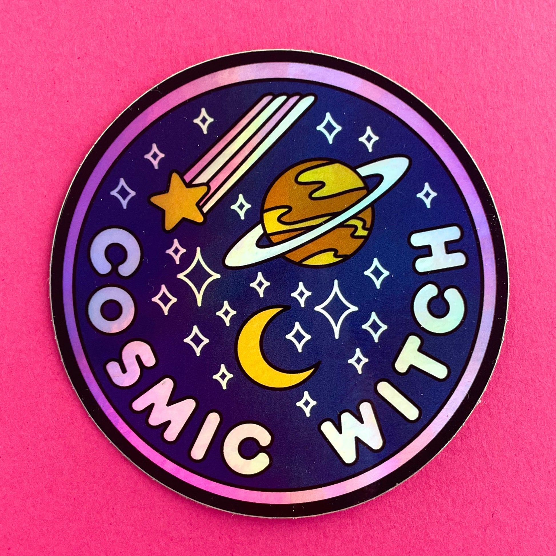 A circular holographic sticker. It has a hot pink border and a dark blue background with light pink words that read "Cosmic Witch". Depicted above the words are a crescent moon, sparkle stars, a shooting star, and the planet Saturn. The sticker is on a hot pink background.