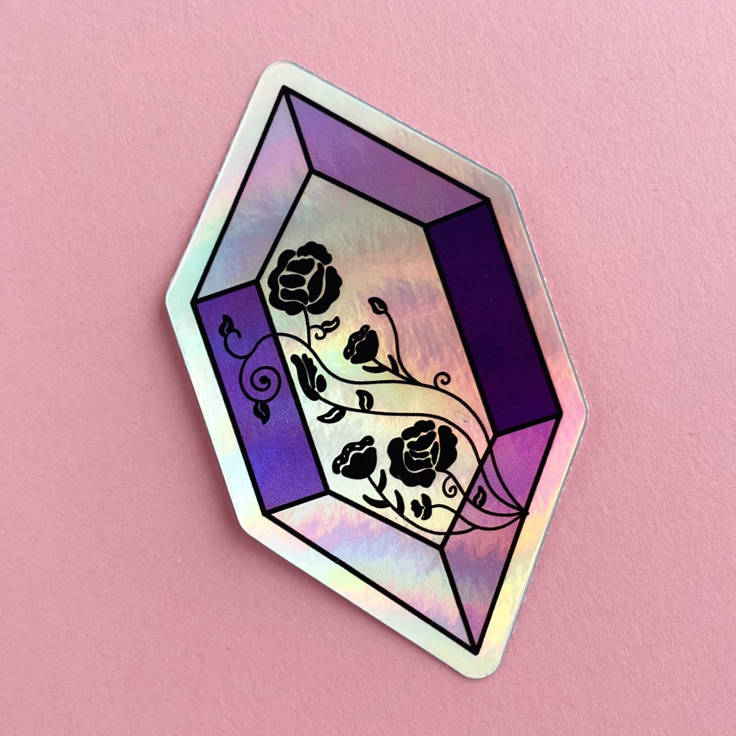 A holographic sticker in the shape of an elongated rectangle. It is various shades of purple and has roses in black line art across the face. The sticker is on a pink background.