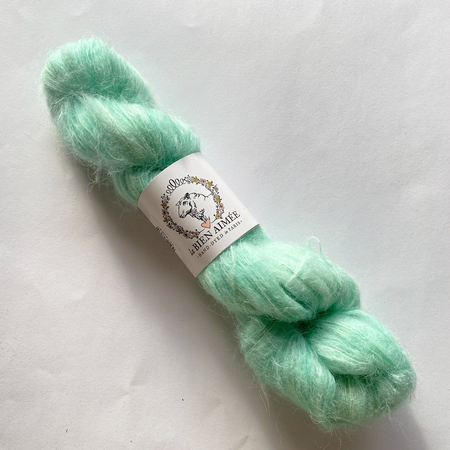 An image of a skein of lace weight yarn in a bright aqua green color with a white ‘La Bien Aimee’ label against a white background.