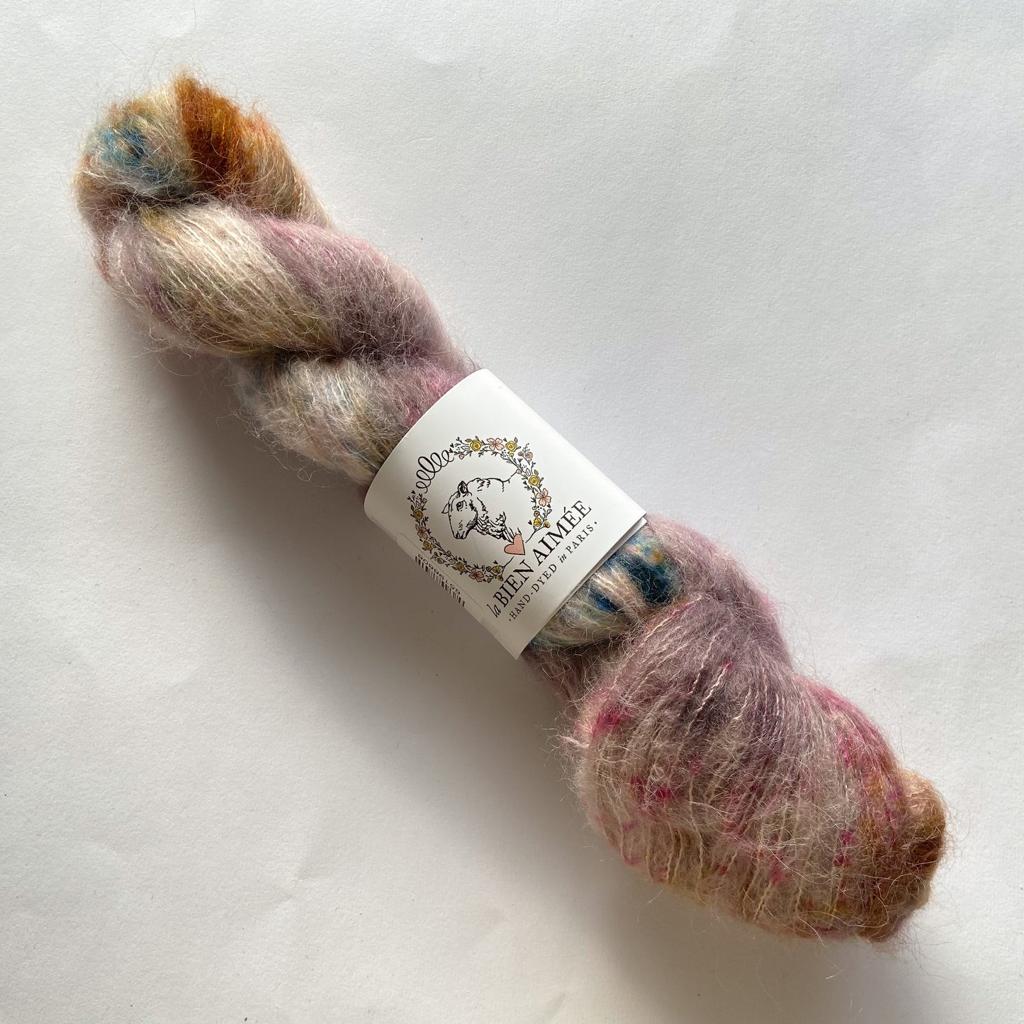 An image of a skein of La Bien Aimee lace weight yarn in the colors mauve, teal, rust orange, and cream with a white ‘La Bien Aimee’ label against a white background.