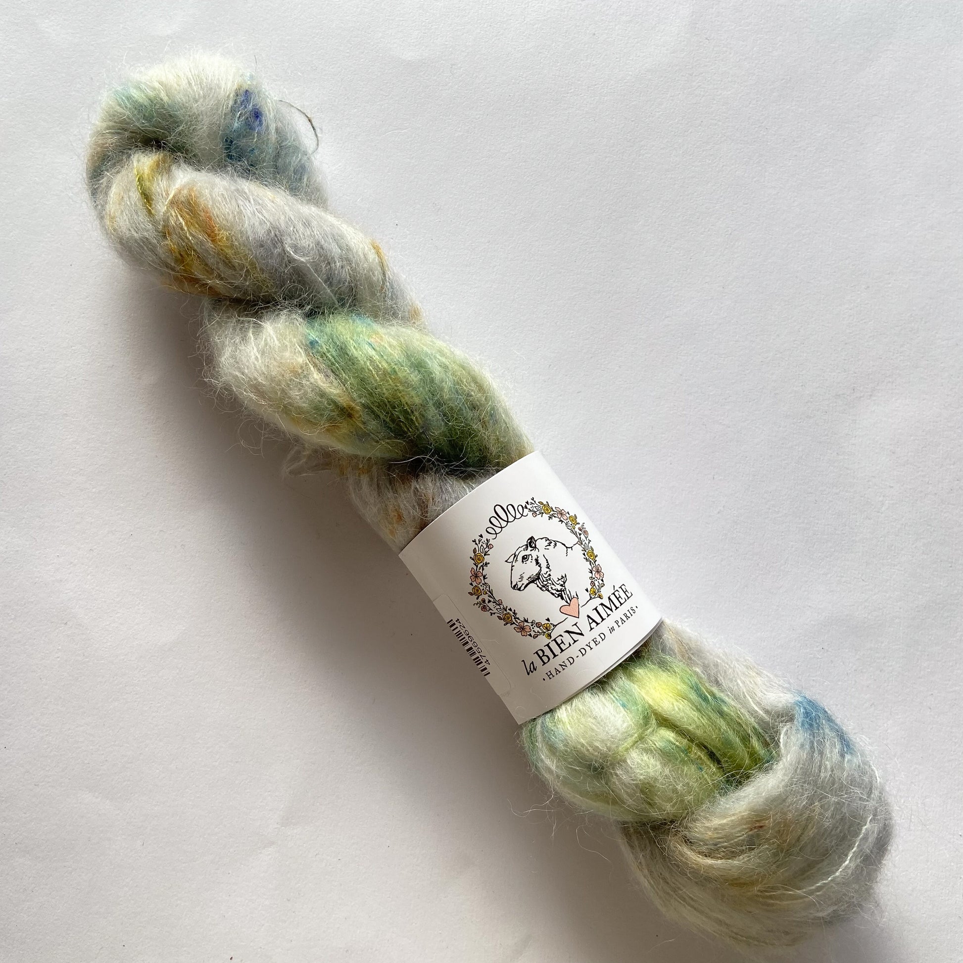 An image of a skein of La Bien Aimee lace weight yarn in the colors gray, green, blue, and yellow with a white ‘La Bien Aimee’ label against a white background.