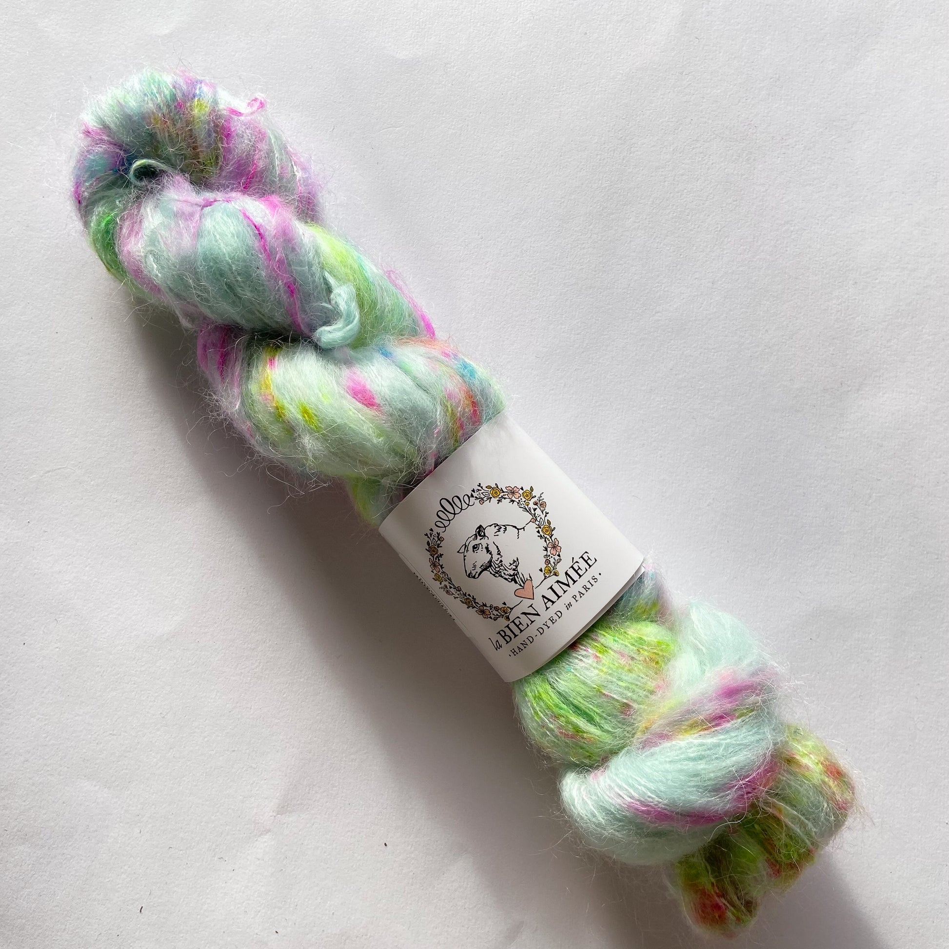An image of a skein of lace weight yarn in a pale blue base with speckles of neon green and pink with a white ‘La Bien Aimee’ label against a white background.