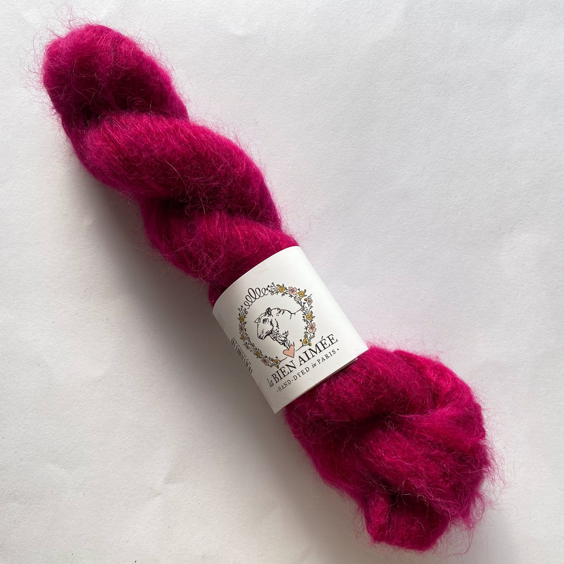 An image of a skein of lace weight yarn in a vibrant fuscia color with a white ‘La Bien Aimee’ label against a white background.