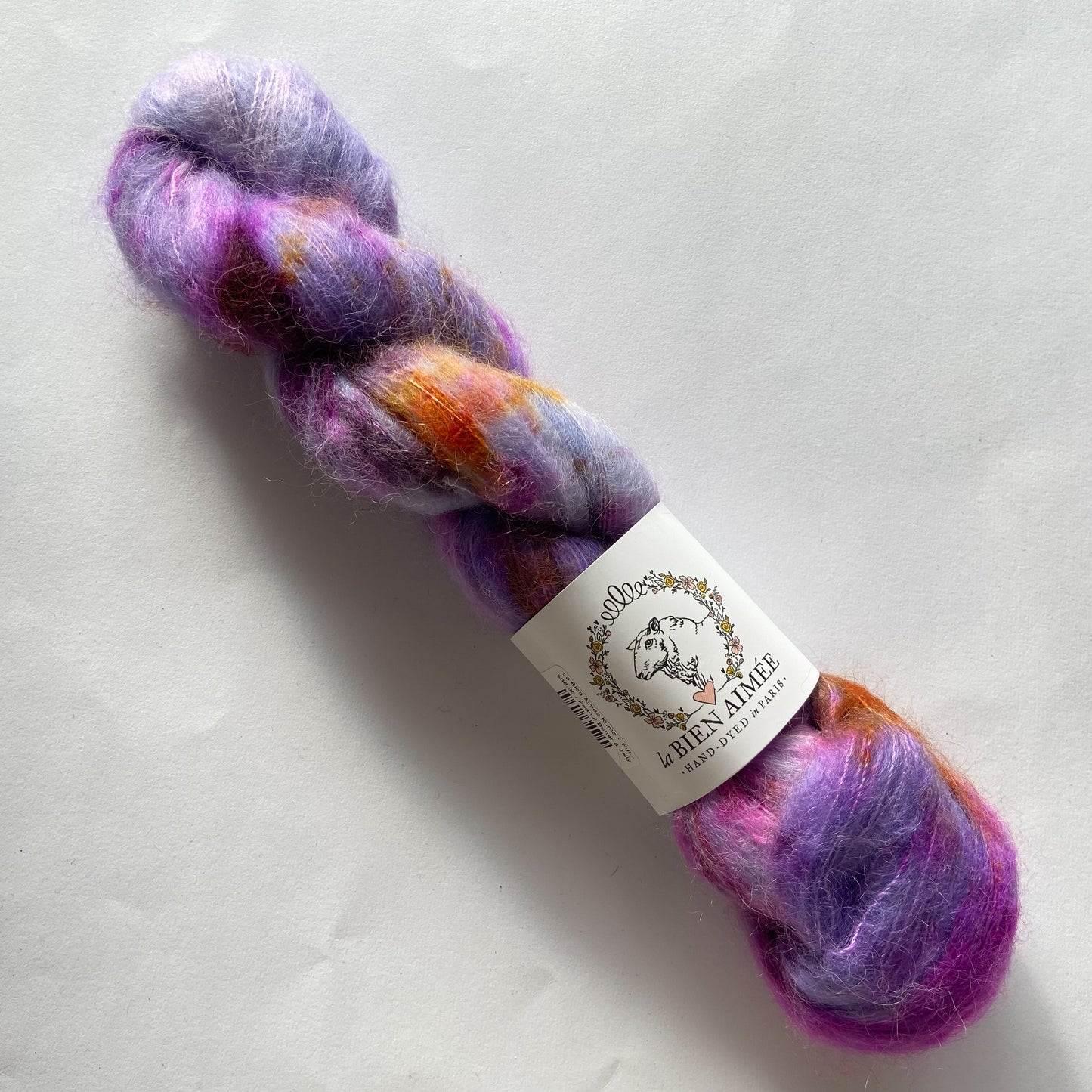 An image of a skein of lace weight yarn in shades of purple, magenta, and orange with a white ‘La Bien Aimee’ label against a white background.