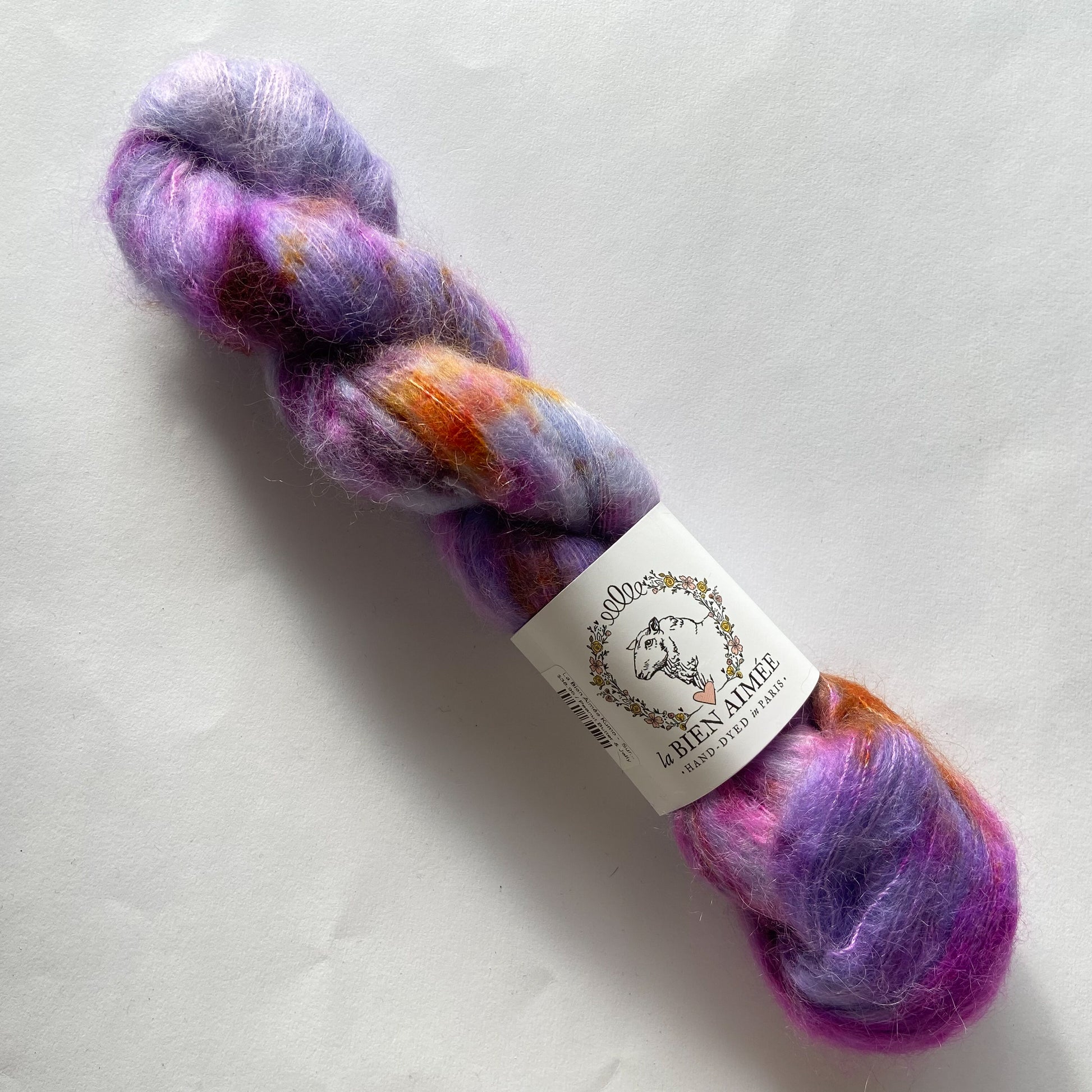 An image of a skein of lace weight yarn in shades of purple, magenta, and orange with a white ‘La Bien Aimee’ label against a white background.