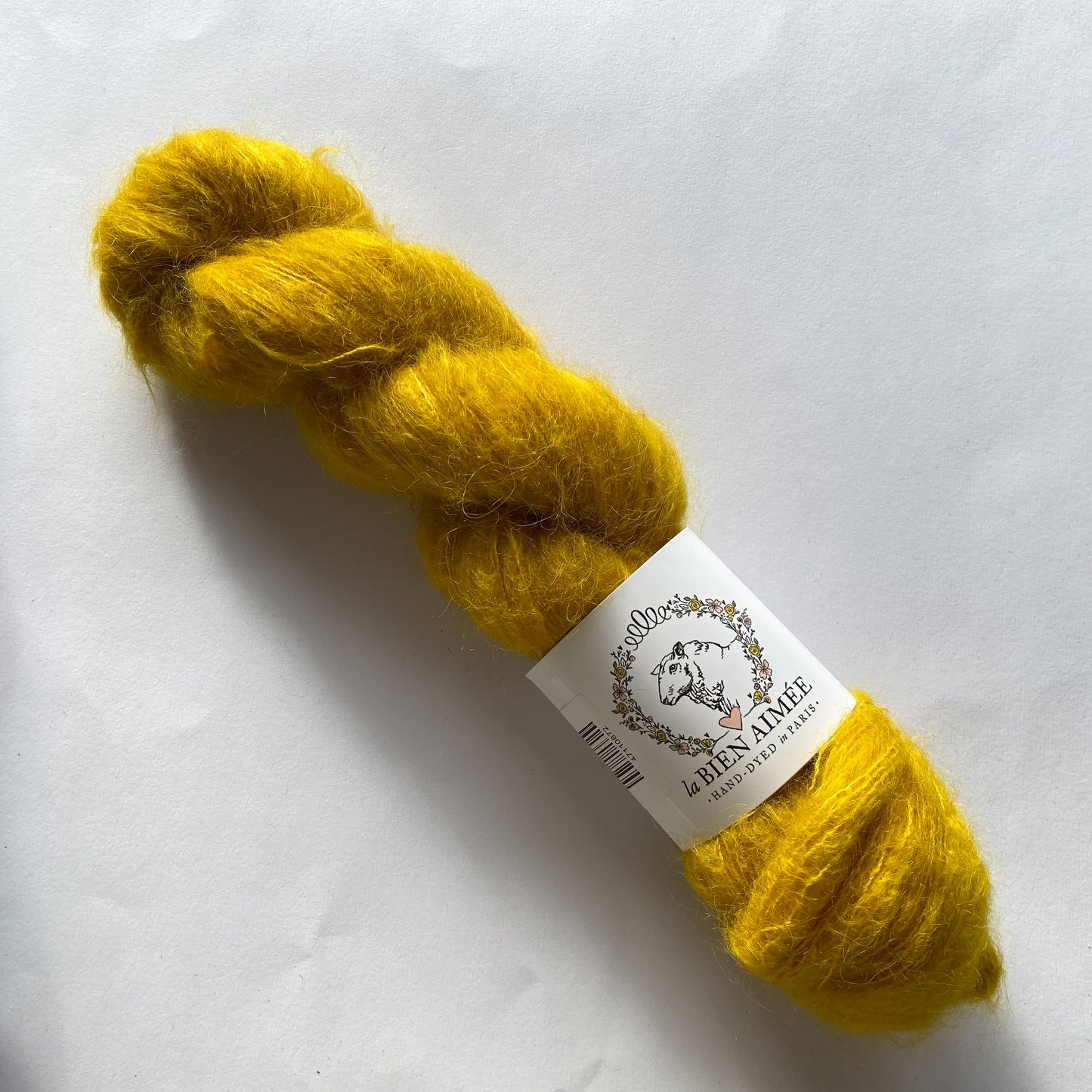 An image of a skein of lace weight yarn in a vibrant mustard yellow color with a white ‘La Bien Aimee’ label against a white background.