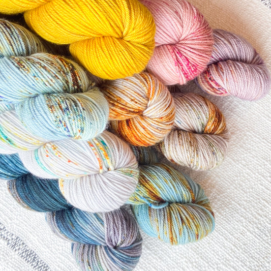 An image of multiple skeins of La Bien Aimee DK weight yarn in a variety of colors against a canvas background.